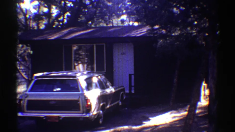 Wooded cabin exterior with a parked station wagon, seen through high-contrast 8mm home-movie exposure.