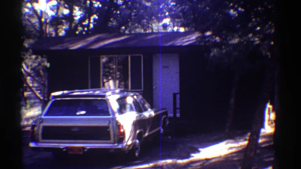 1969 Arizona 8mm frame showing a large station wagon parked near a small dark-sided house in deep tree shade.