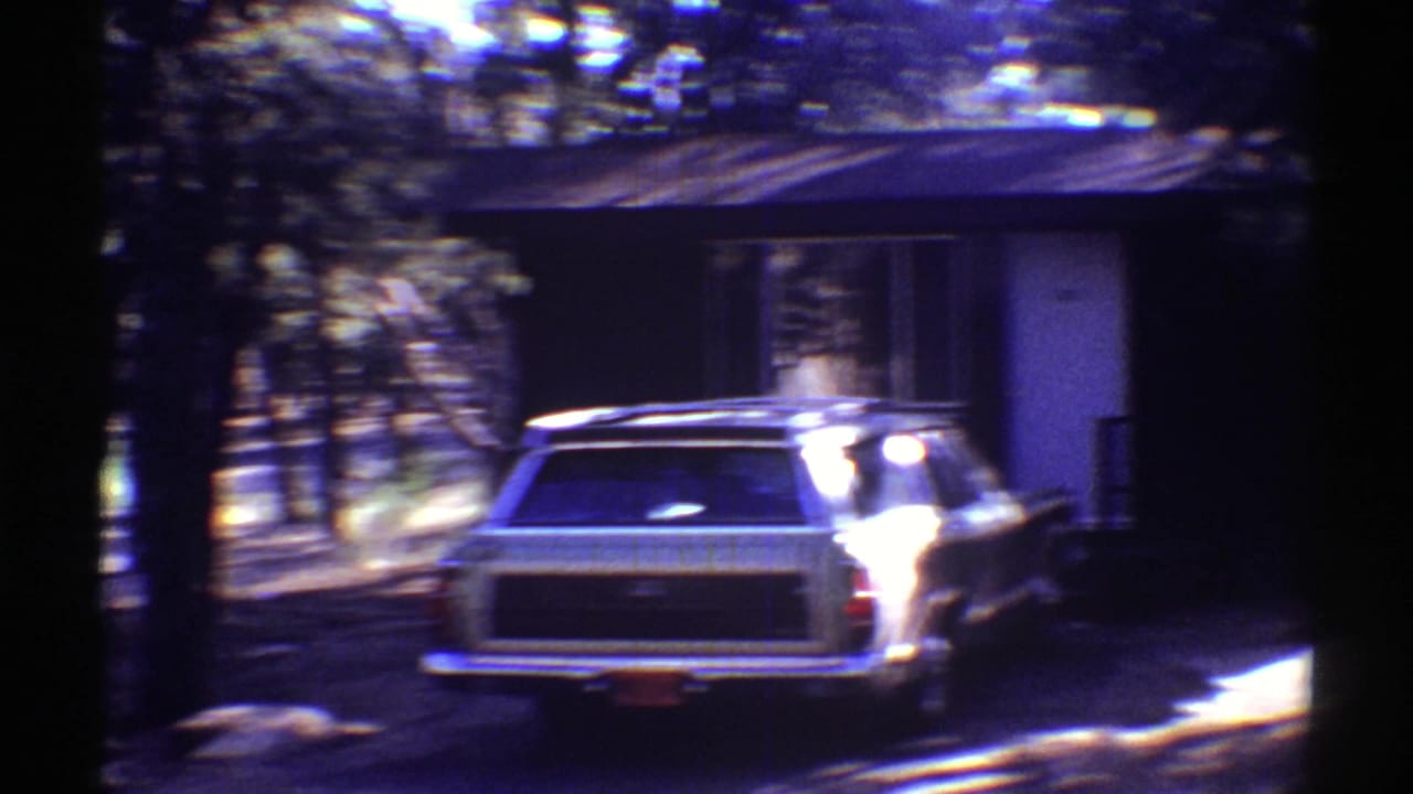 Rear view of a vintage station wagon near a modest house, with trees and bright sun patches in the Arizona setting.