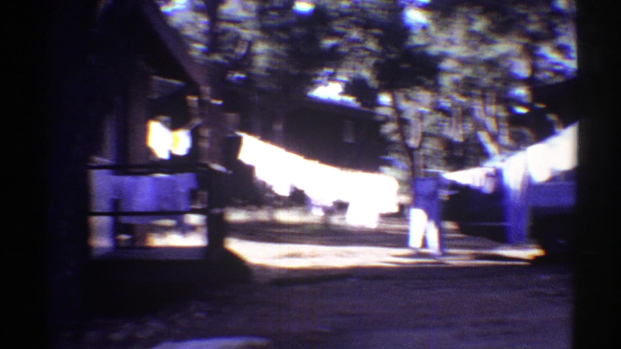 1969 Arizona home-movie frame showing laundry on a line beside trees, a driveway, and a parked car in deep shade.