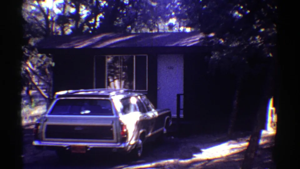 A station wagon sits in the driveway of a small Arizona house, with trees casting shadows across the 1969 home-movie frame.