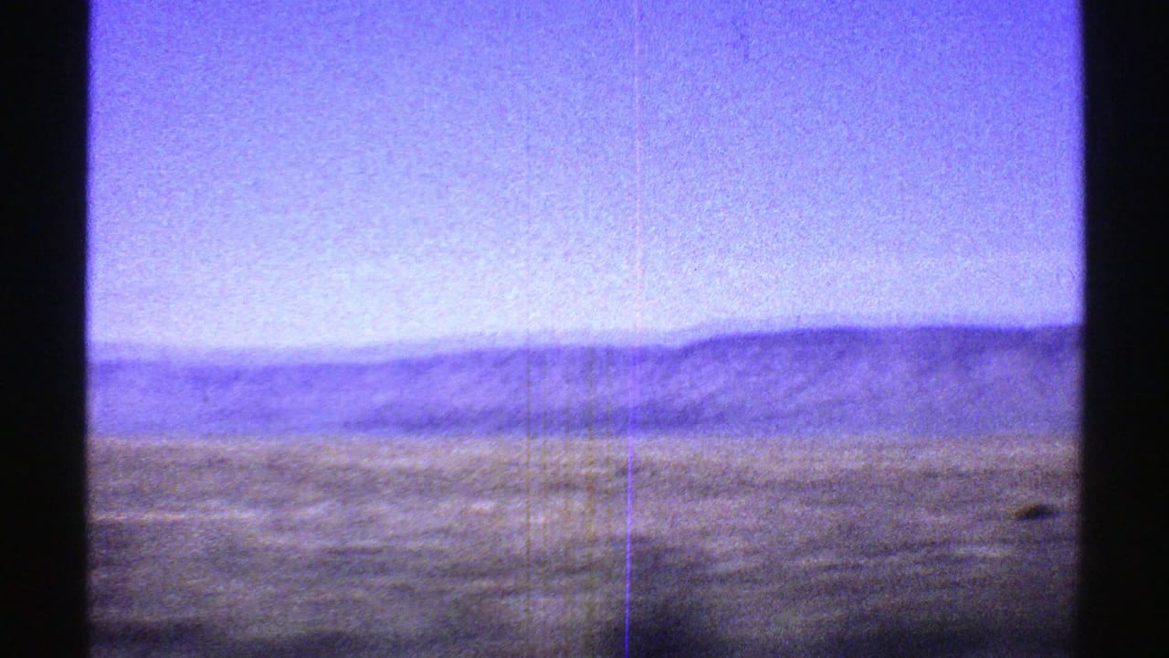 Distant mountain ridge beyond open grassland under a pale sky, seen through film-frame borders.