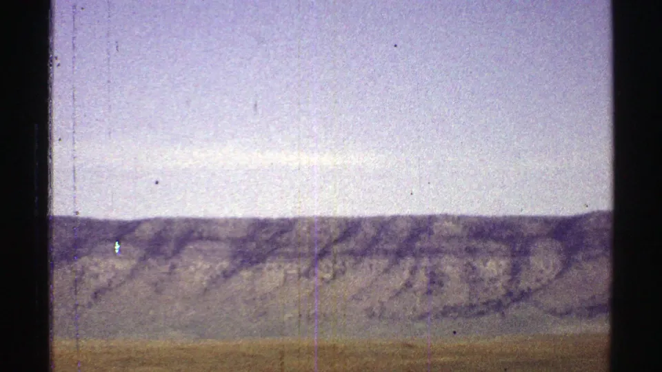 1969 Arizona 8mm frame showing scrubby desert terrain, a distant ridge, pale sky, grain, and vertical wear marks.