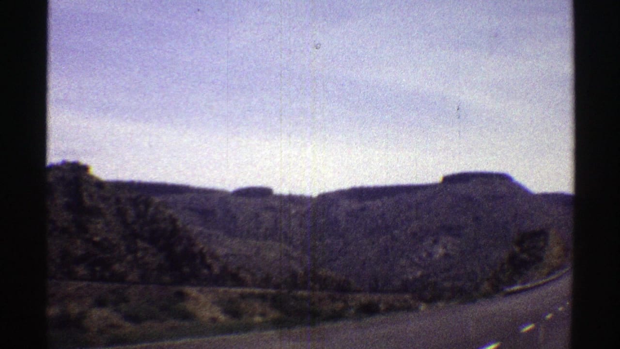 1969 Arizona home-movie frame showing a paved road curving past scrub and rocky hills beneath a pale sky.