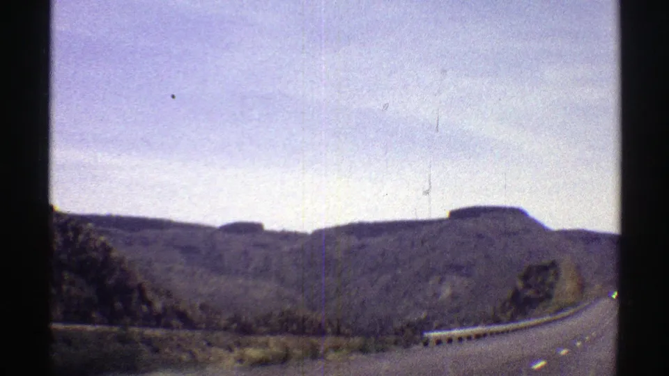 Arizona road scene from a moving vehicle, with guardrail, rocky slopes, and muted 8mm color.