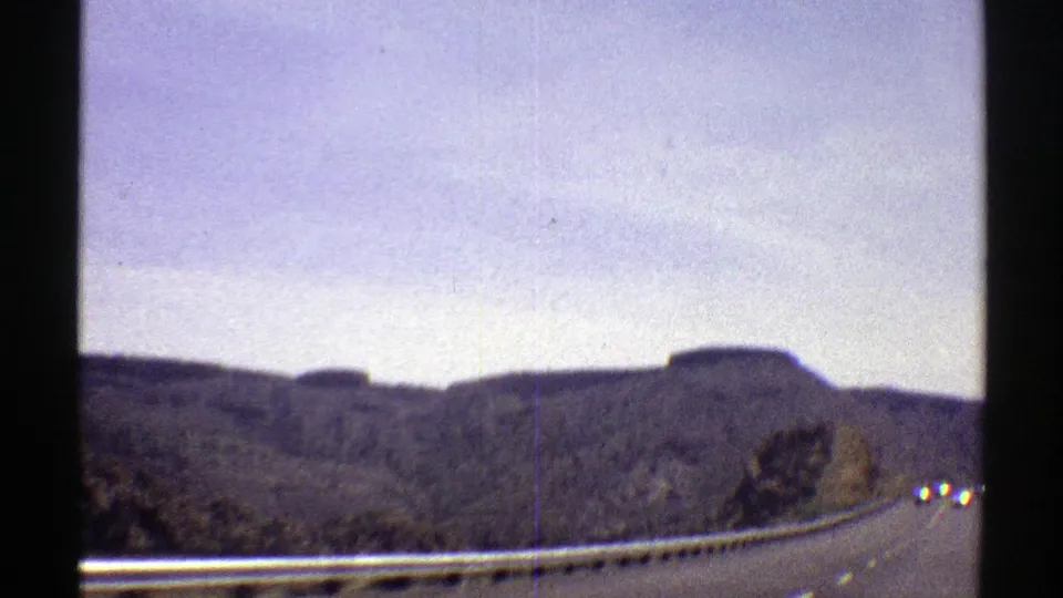 1969 Arizona driving view with a highway guardrail, distant vehicles, rocky terrain, and soft 8mm grain.