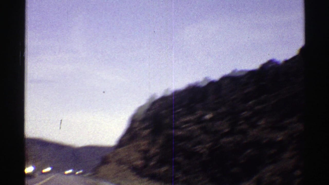 Passing rocky hillside under pale blue sky with dark film borders.