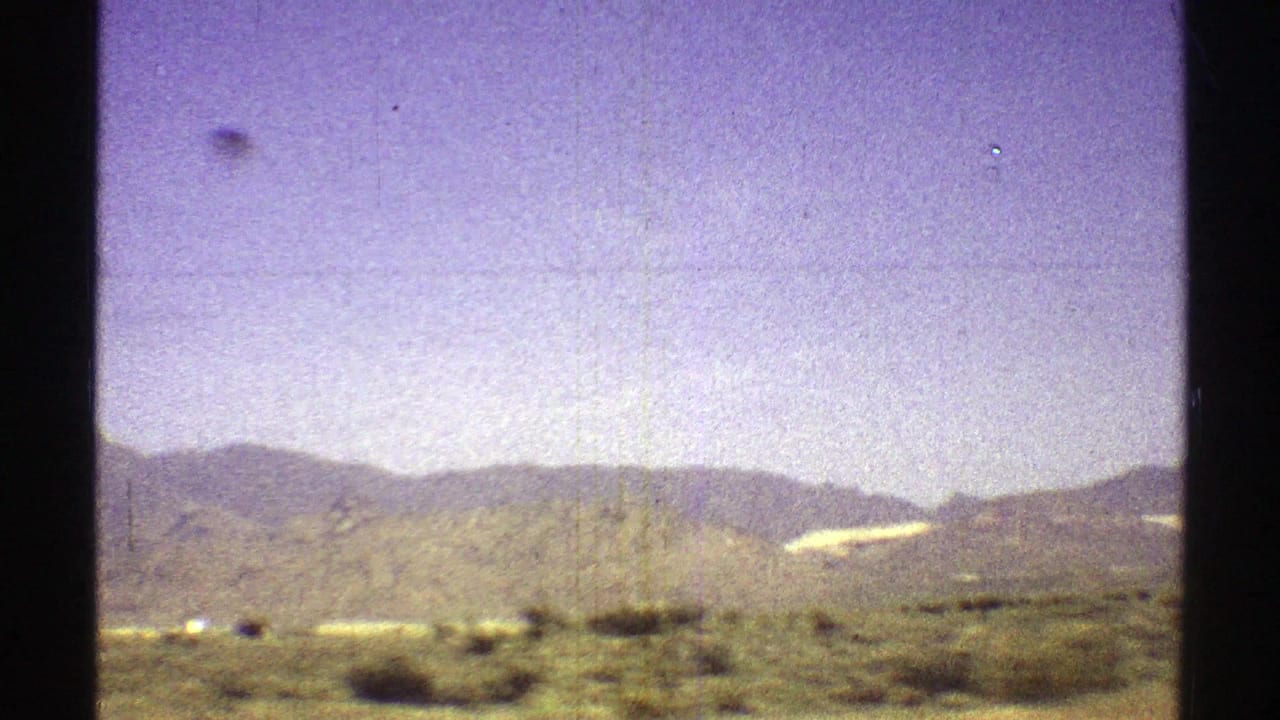 Desert shrubs and low mountains seen from a moving vehicle under a pale sky.