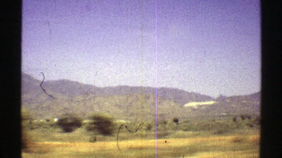 1969 Arizona desert driving frame with hazy mountains, scattered brush, pale sky, and visible 8mm scratches.
