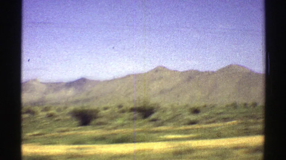 Arizona 1969 landscape frame showing a mountain ridge beyond scrubby desert ground, with grain and scratches from 8mm film.