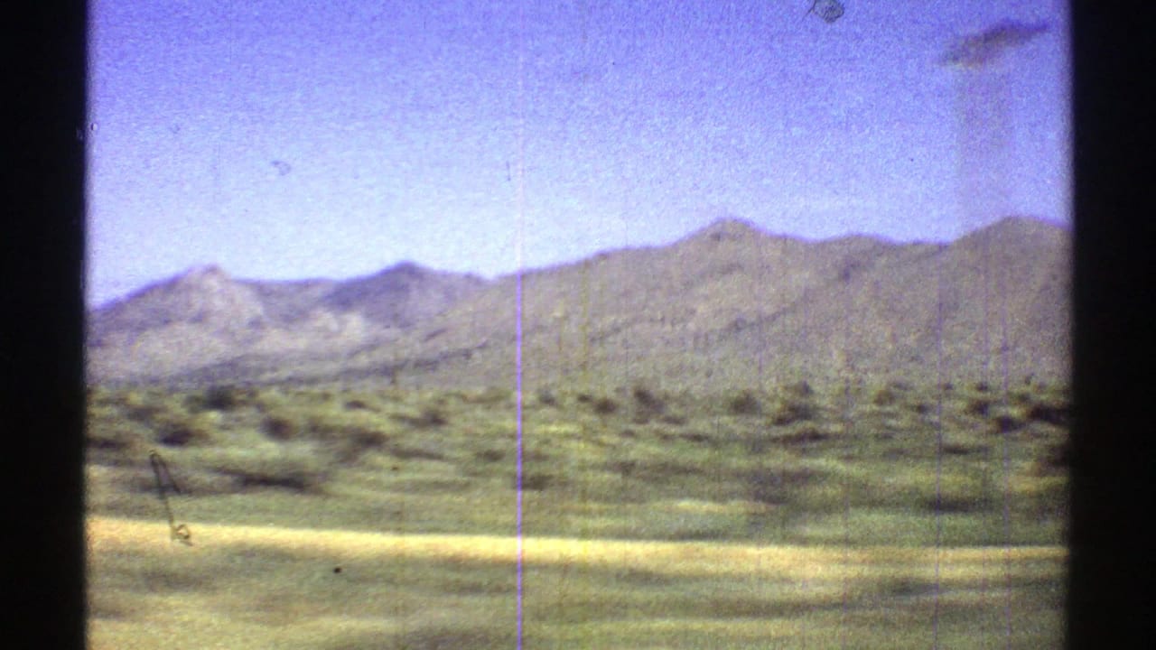 1969 Arizona 8mm frame showing green desert vegetation, pale roadside ground, and distant mountain ridges beneath a bright blue sky.