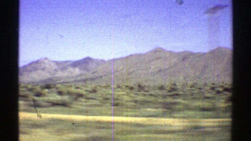 1969 Arizona 8mm frame showing green desert vegetation, pale roadside ground, and distant mountain ridges beneath a bright blue sky.