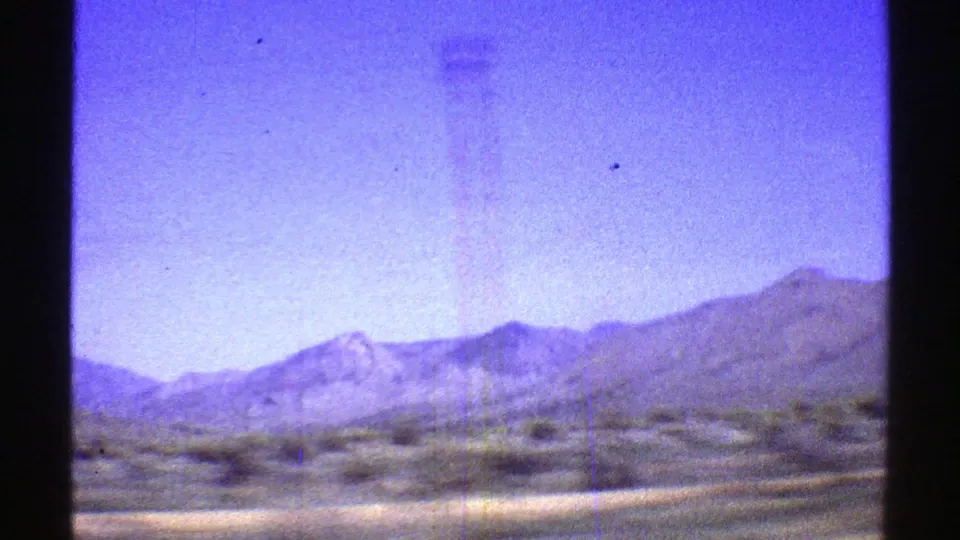 1969 Arizona 8mm frame with motion-blurred roadside desert shrubs, distant mountains, blue sky, and visible film wear from a moving vantage point.