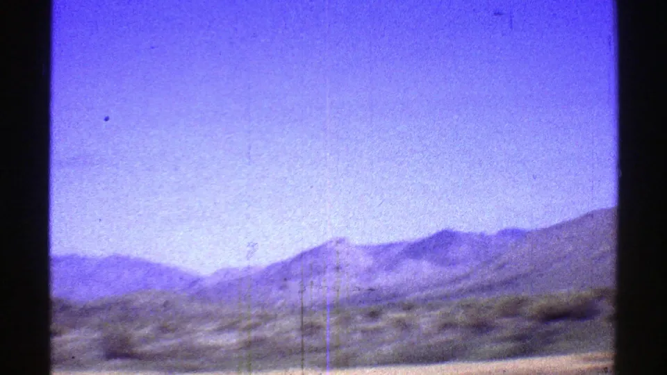 1969 Arizona 8mm frame showing distant desert mountains, low vegetation, several thin utility poles, and a bright blue sky with visible film grain.