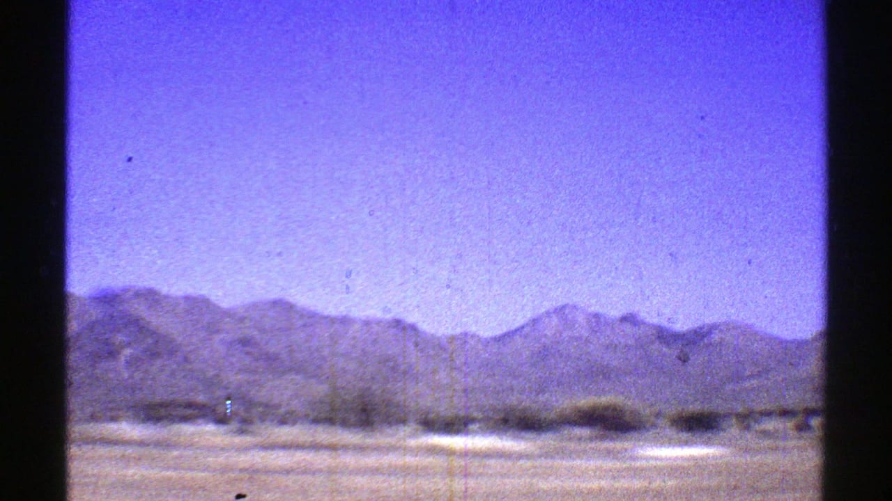 Arizona desert mountains appear beyond a pale roadside strip, filmed on 8mm from inside a moving vehicle in 1969.