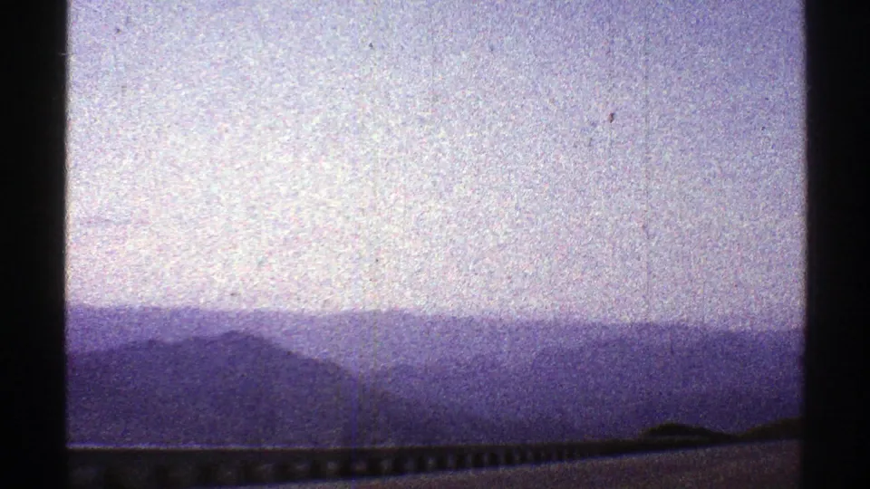 A purple-shifted 8mm view of Arizona mountain ridges, roadside guardrail, and pale sky from a moving car in 1969.