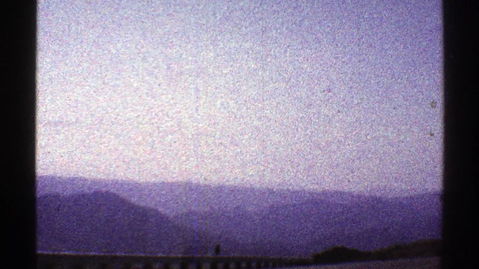 Final 1969 Arizona frame with layered mountain silhouettes, hazy sky, guardrail, and aged 8mm grain visible across the scene.