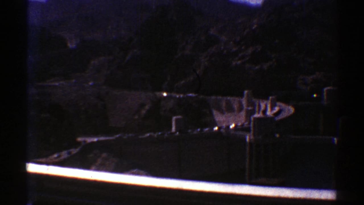 Visitor-shot 8mm view of a concrete dam set against dark Arizona canyon walls, partly framed by a bright rail.