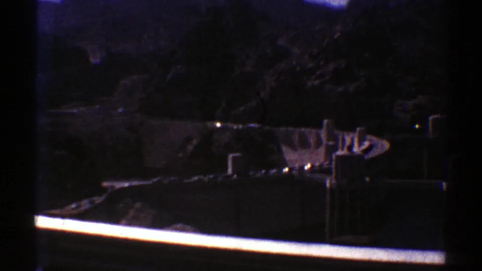 Visitor-shot 8mm view of a concrete dam set against dark Arizona canyon walls, partly framed by a bright rail.