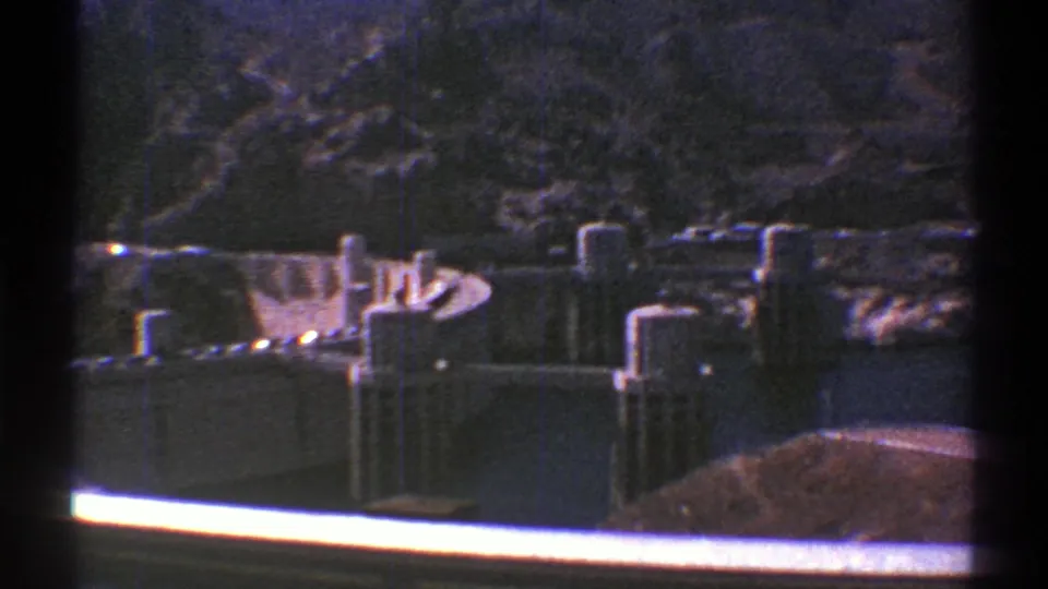 Late-1960s 8mm view of an Arizona canyon dam, with concrete towers, shadowed rock, and a bright rail crossing the foreground.