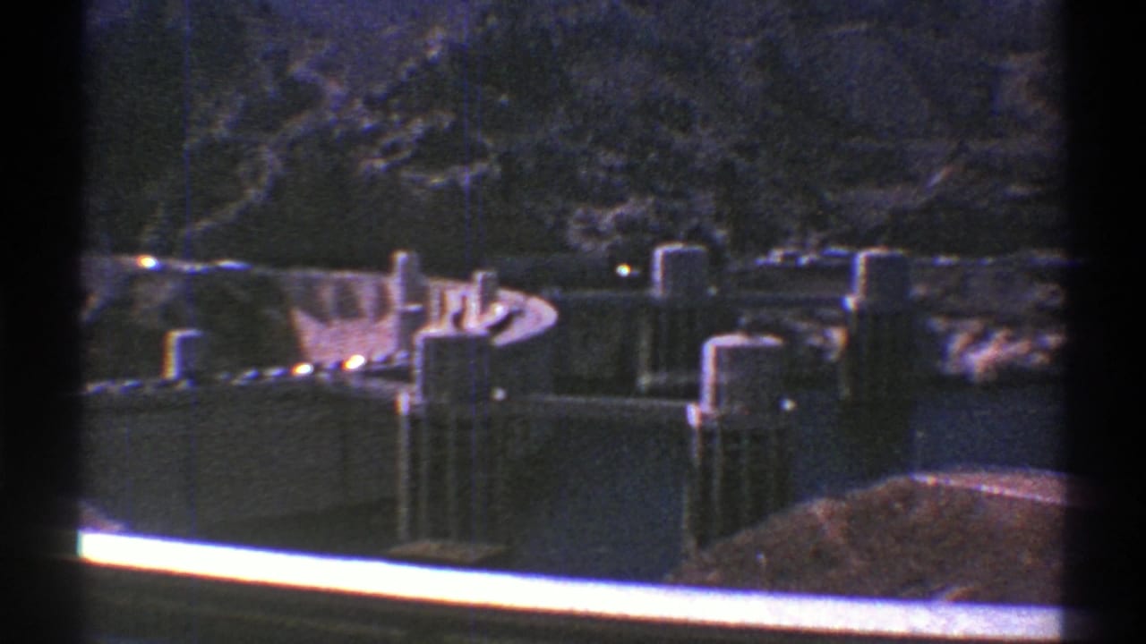1969 Arizona 8mm frame showing a concrete dam, spillway structures, towers, reservoir water, and rugged mountain slopes.