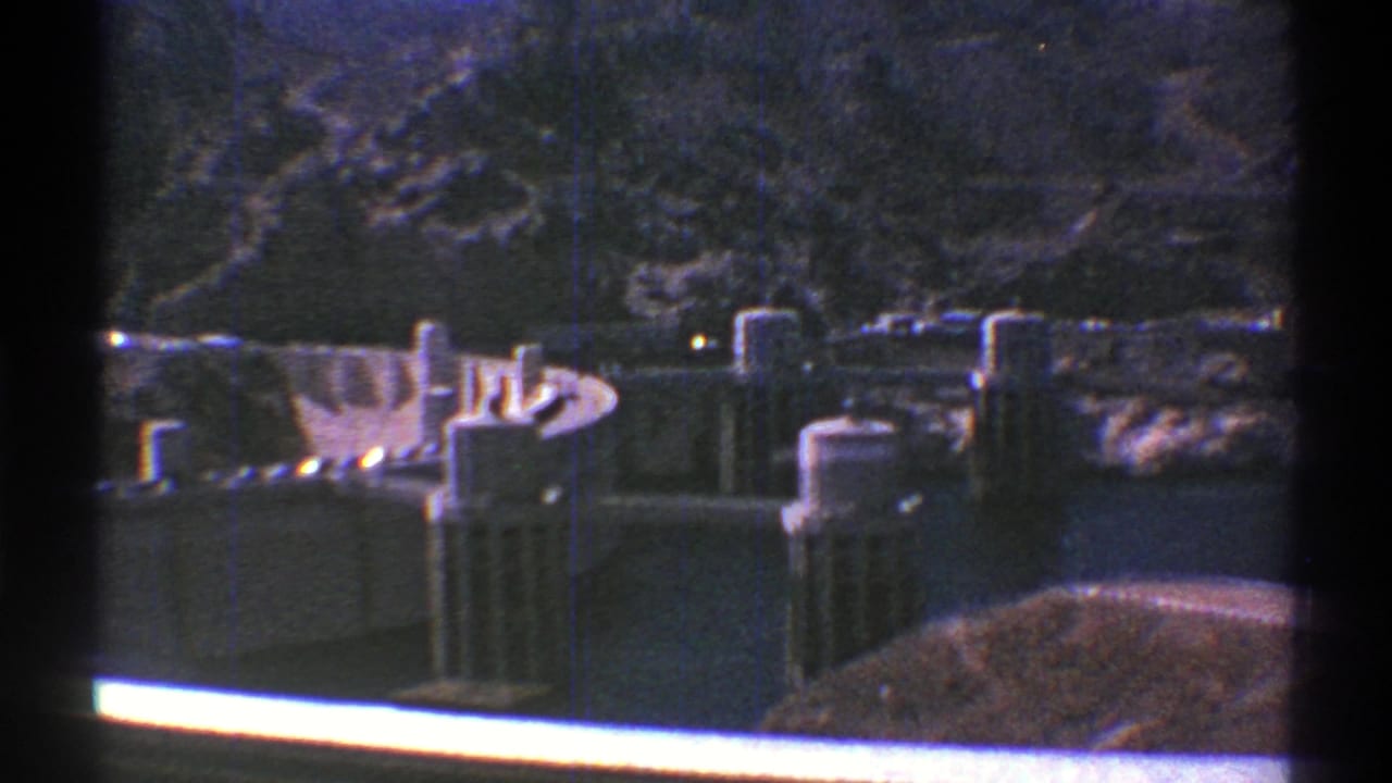 1969 Arizona home-movie frame of a dam and reservoir, with concrete towers, spillway forms, and steep rocky terrain in the background.