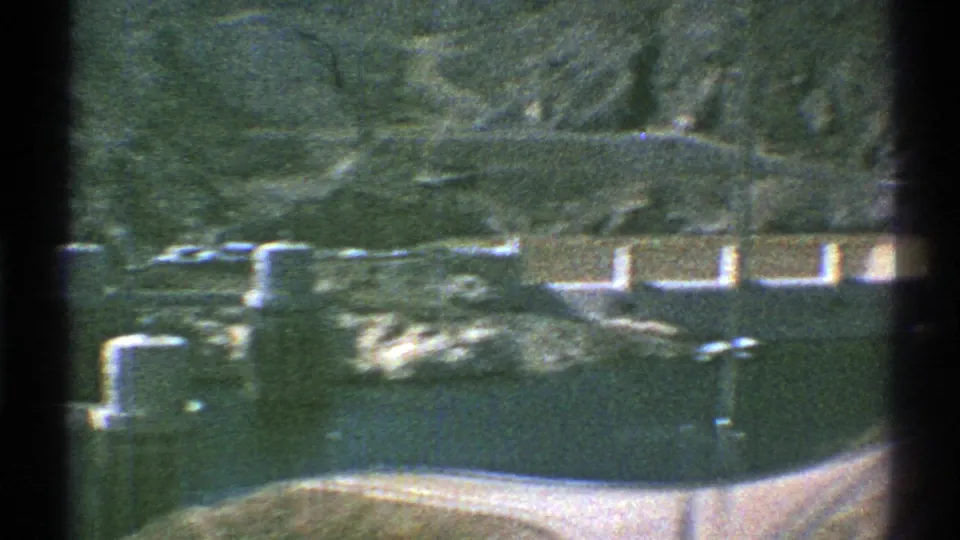 Faded 8mm frame from Arizona in 1969 showing the dam wall, reservoir, low towers, shoreline brush, and rocky slopes.