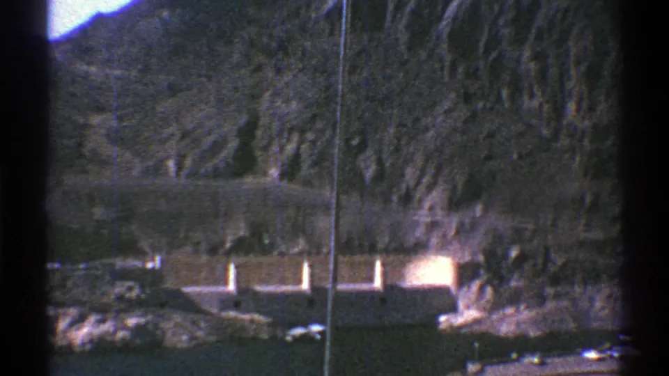 1969 Arizona 8mm frame showing a concrete dam across dark water, backed by steep desert mountains and a vertical film scratch.