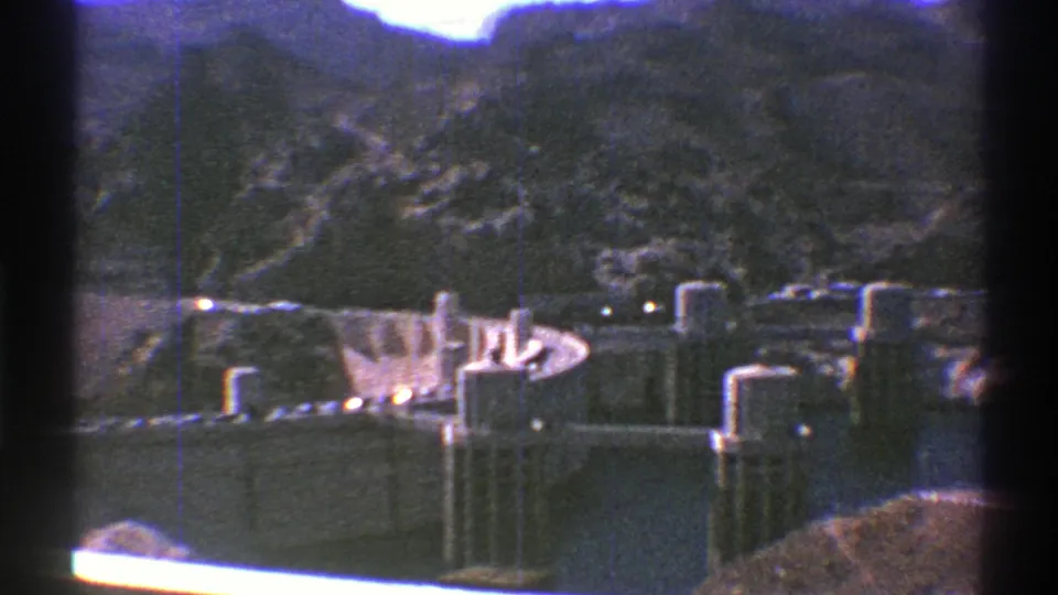 Late-1960s 8mm view of Hoover Dam from above, with concrete dam structures, power buildings, water, and rocky Arizona landscape.