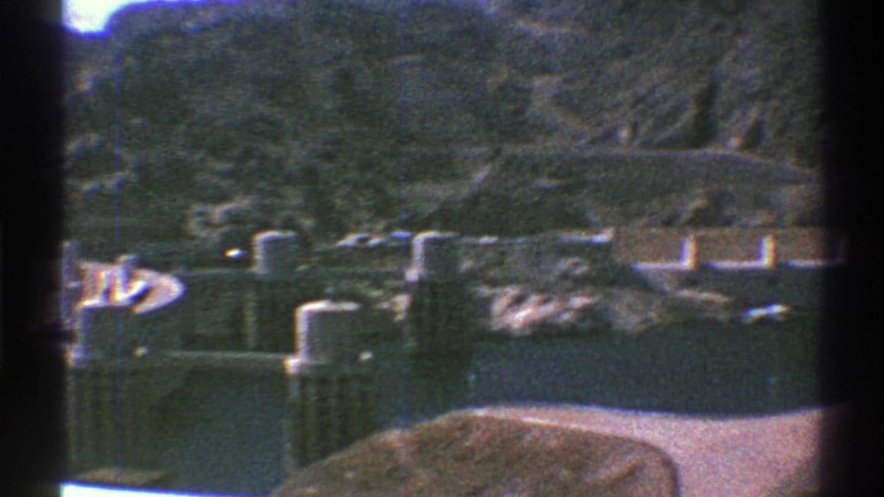 1969 Arizona home-movie frame showing a dam, reservoir edge, and rugged hills in bright daylight.