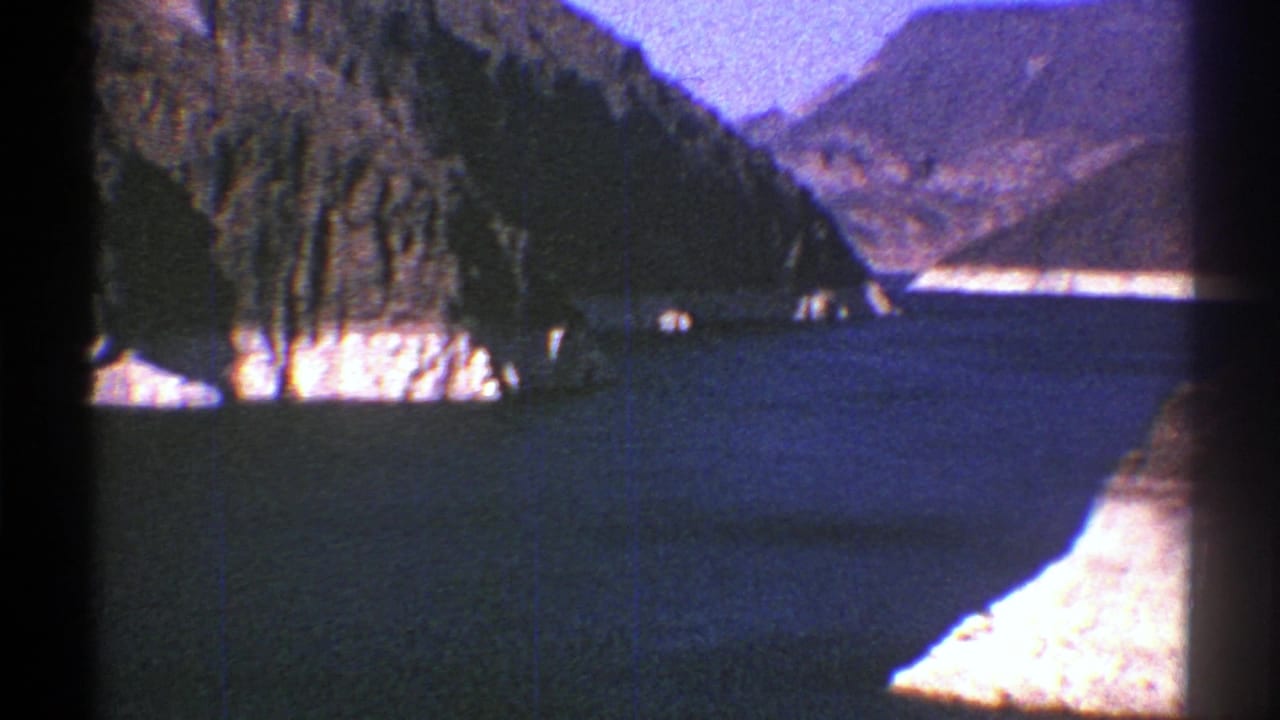 1969 Arizona 8mm frame showing deep blue water, pale cliff faces, and rugged mountain slopes under a clear sky.