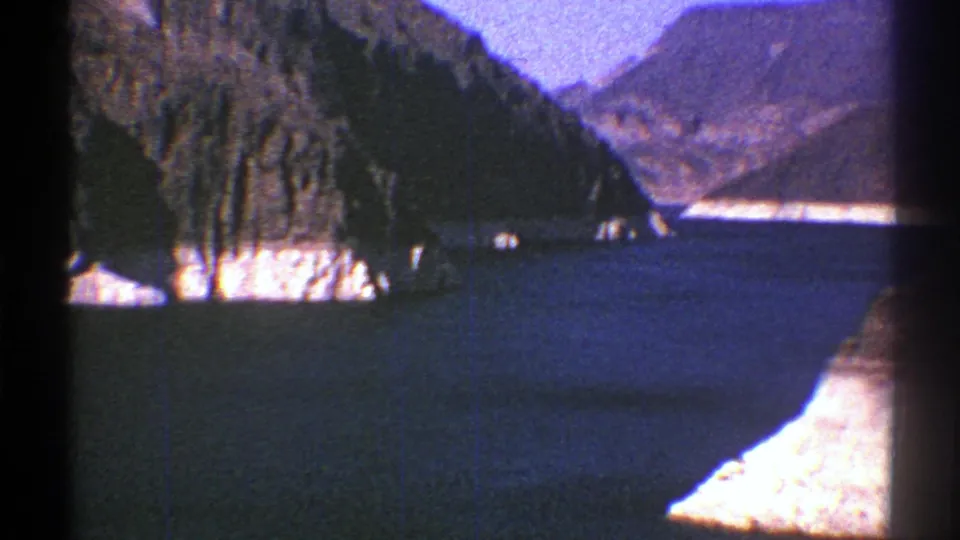 1969 Arizona 8mm frame showing deep blue water, pale cliff faces, and rugged mountain slopes under a clear sky.