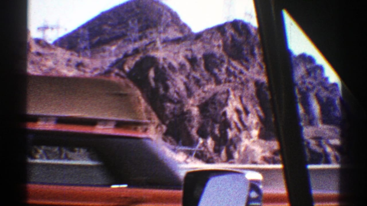 1969 Arizona road footage seen through a car window, with rocky desert mountains, highway edge, and part of another vehicle visible.