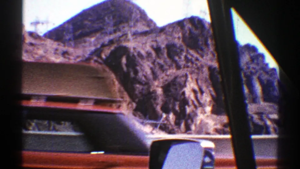 1969 Arizona road footage seen through a car window, with rocky desert mountains, highway edge, and part of another vehicle visible.