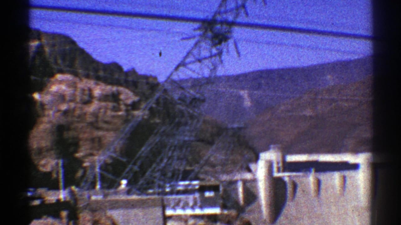 Concrete dam and power transmission tower set against rocky brown mountains.