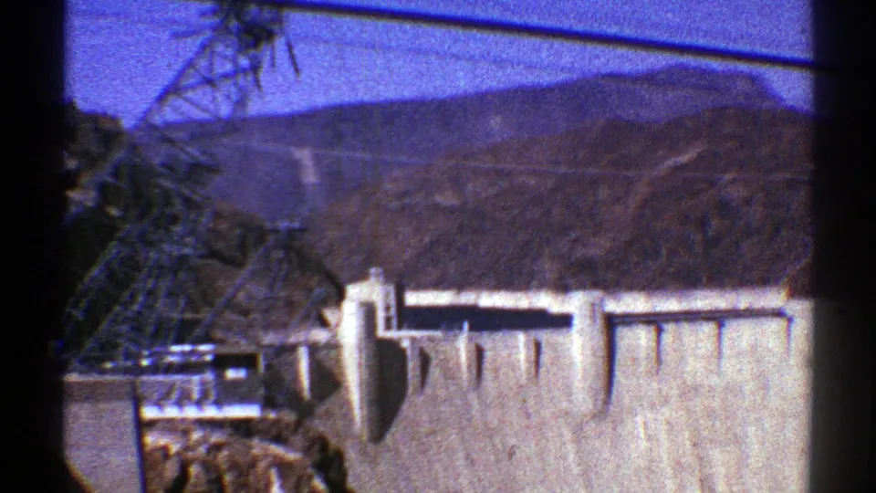 1969 Arizona 8mm frame showing the dam face, a nearby transmission tower, overhead wires, and distant desert ridges.