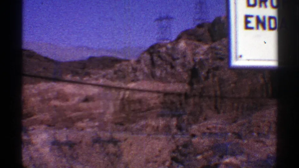 The pan reaches rocky canyon ground and a partial roadside sign near Hoover Dam, with desert hills in the background.