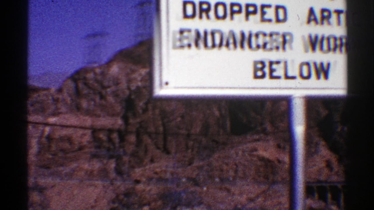 White warning sign in front of rocky terrain near a large dam.