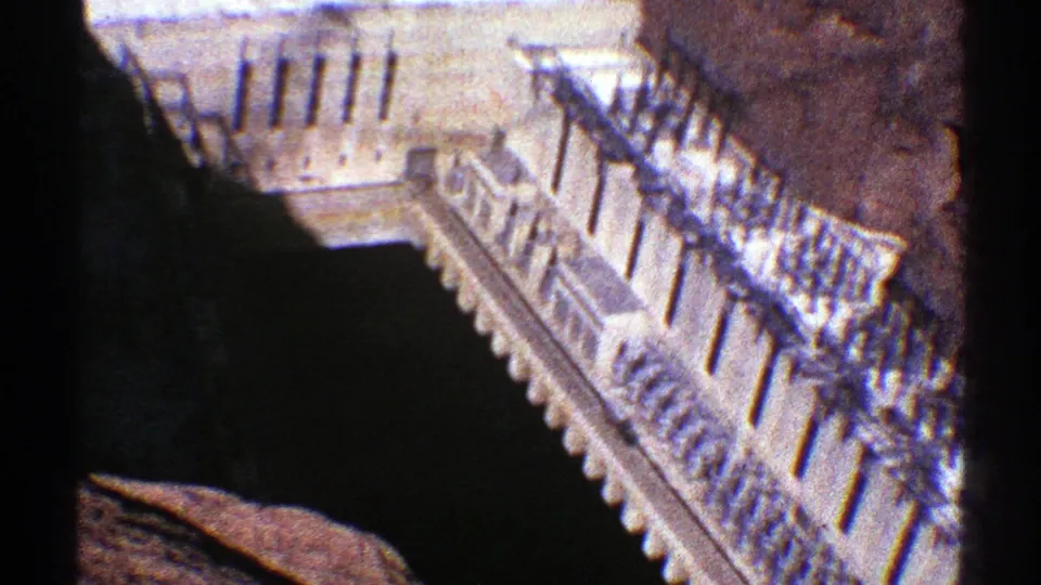 1969 8mm frame of Hoover Dam base structures, showing railings, stairs, concrete surfaces, and heavy canyon shadow.