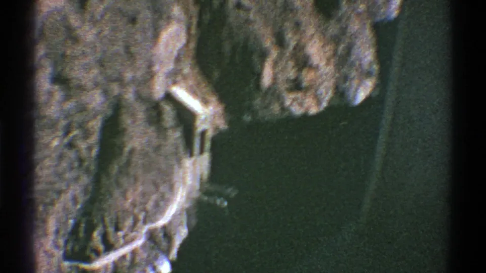 Aerial 8mm frame from Arizona showing rocky terrain, dark water, and a small pale waterside structure or bridge-like feature.