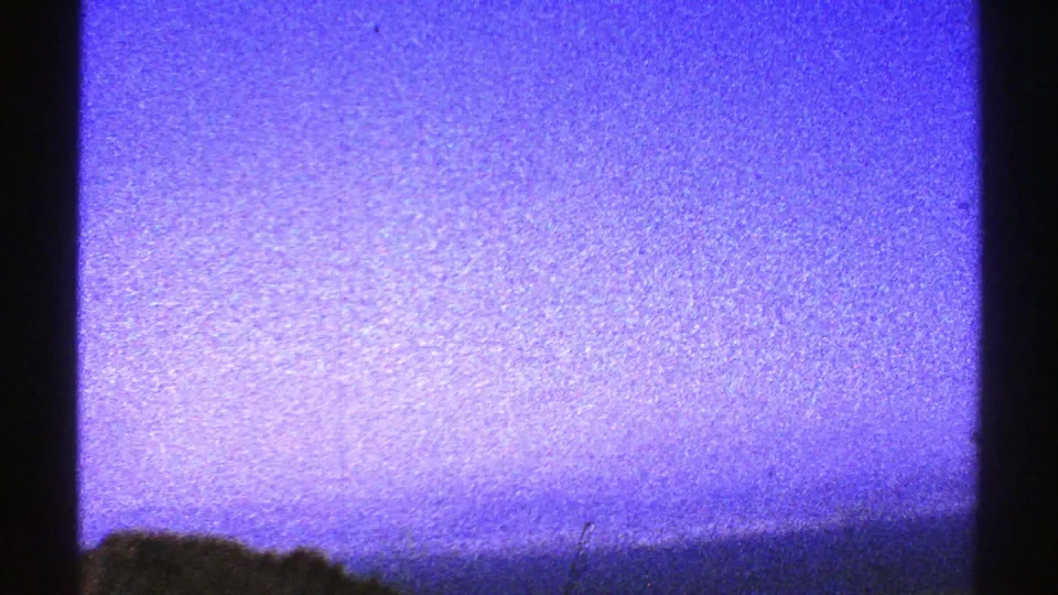 1969 Arizona 8mm frame showing blue-purple sky, faint horizon, low dark foreground shapes, and dense film grain.
