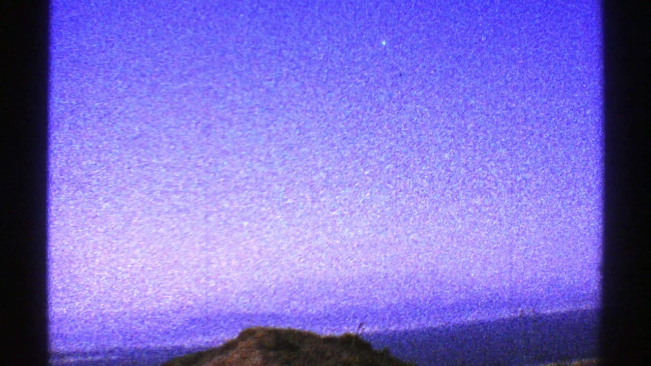 Low Arizona desert hills and hazy horizon appear beneath a purple-blue sky in grainy 8mm footage.