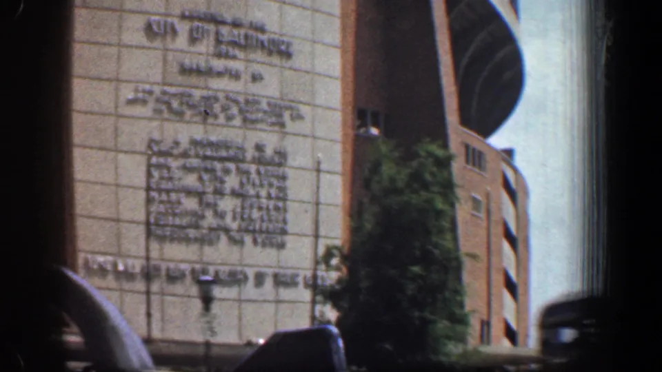 Passenger-shot 1961 Baltimore frame with the building inscription partly readable, a street lamp, tree foliage, and foreground car framing.