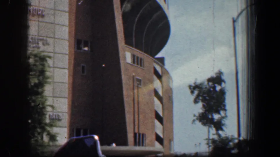 Final 8mm frame from the moving car showing the brick side wall, curved adjacent structure, tree, sky, and dark window framing.
