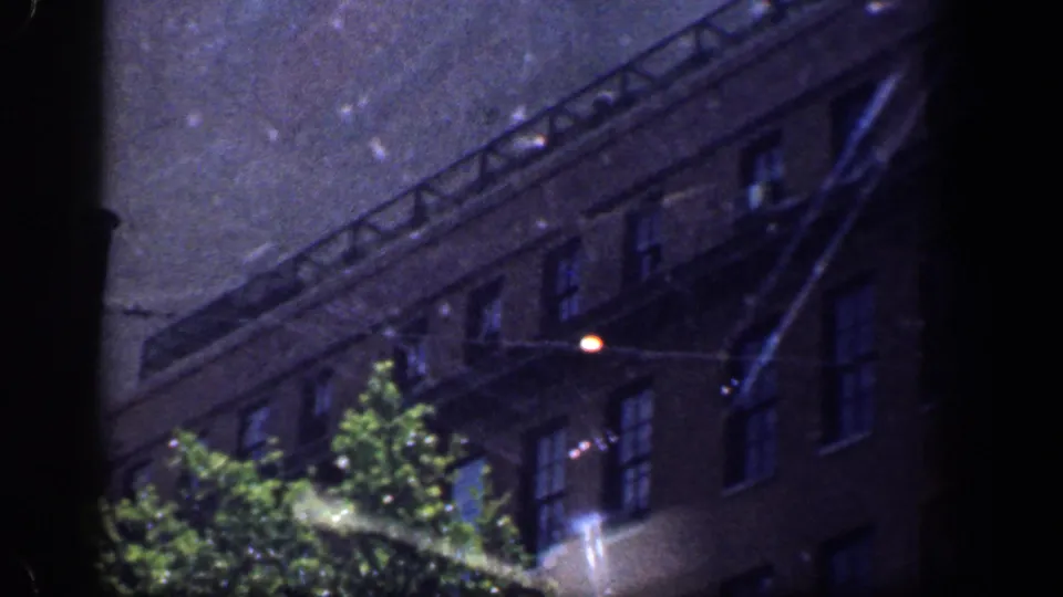 Dark 8mm frame showing a brick building at night with upper windows, leafy trees, streetlight glow, and visible film wear.