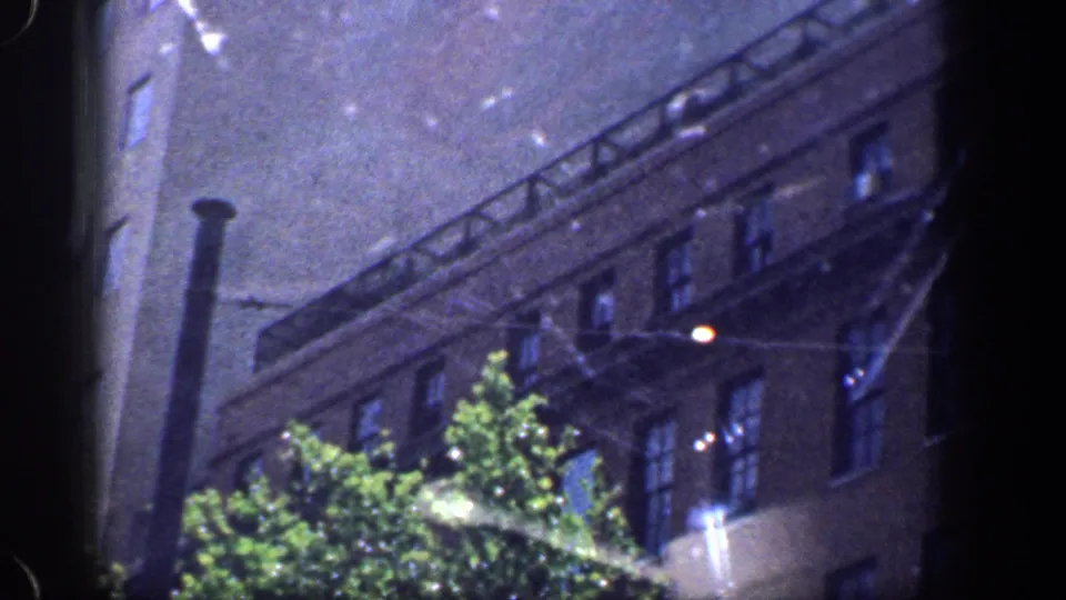 1961 Baltimore night frame with a brick building facade, lit windows or reflections, tree canopy, and a bright streetlight flare.