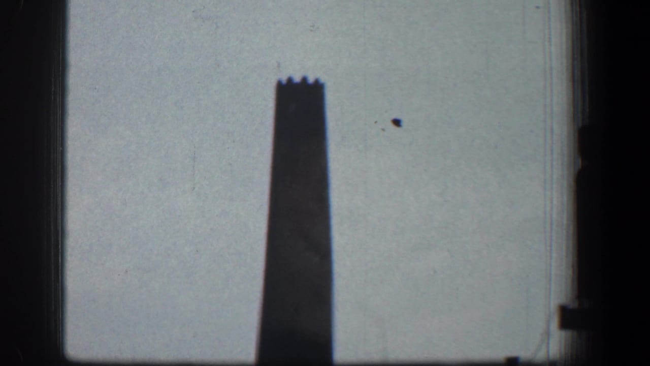 The smokestack remains centered against the pale sky, with dark vertical borders and visible 8mm film texture.