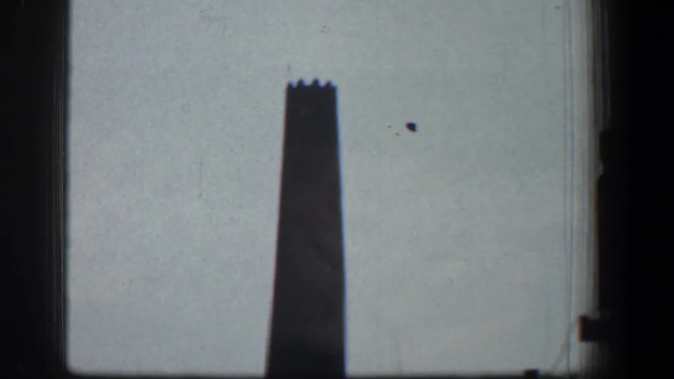 The smokestack remains centered against the pale sky, with dark vertical borders and visible 8mm film texture.