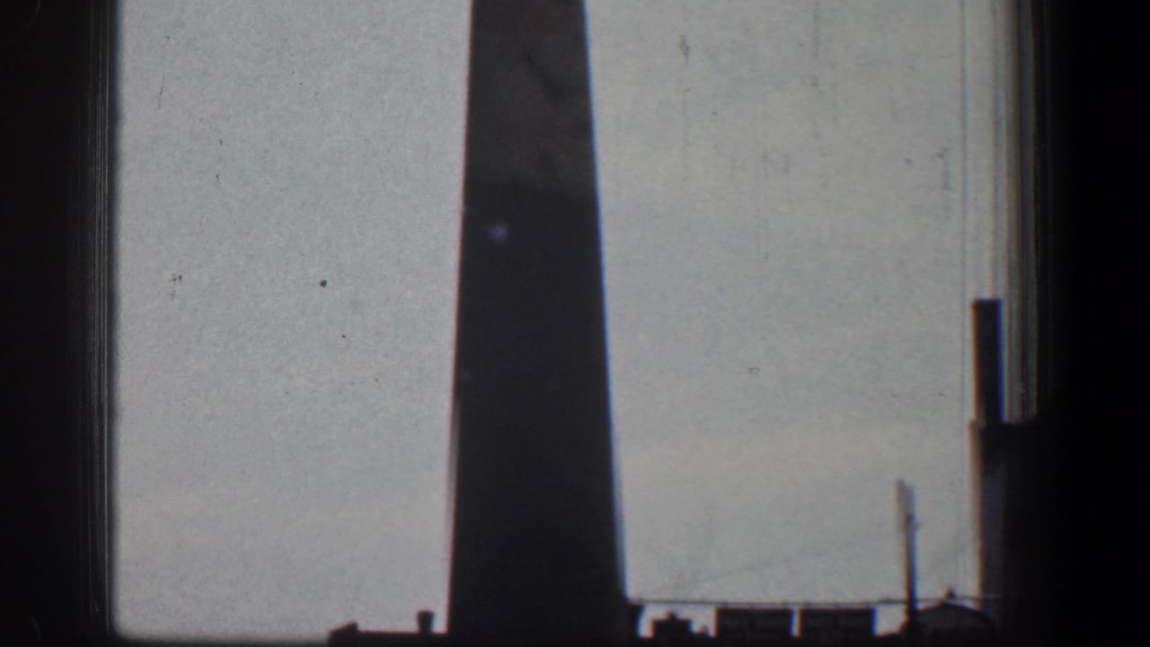 1961 Baltimore home-movie frame showing a tall industrial chimney against a pale sky, with dark gate edges and distant roofline details.