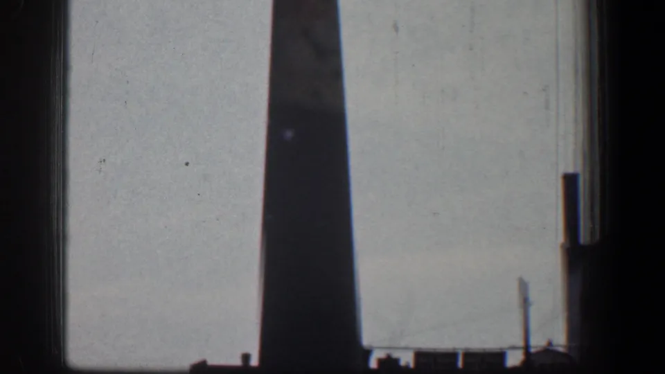 1961 Baltimore home-movie frame showing a tall industrial chimney against a pale sky, with dark gate edges and distant roofline details.